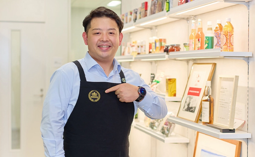 プレミアムエプロンを身に付けて胸元のロゴを指すTさん